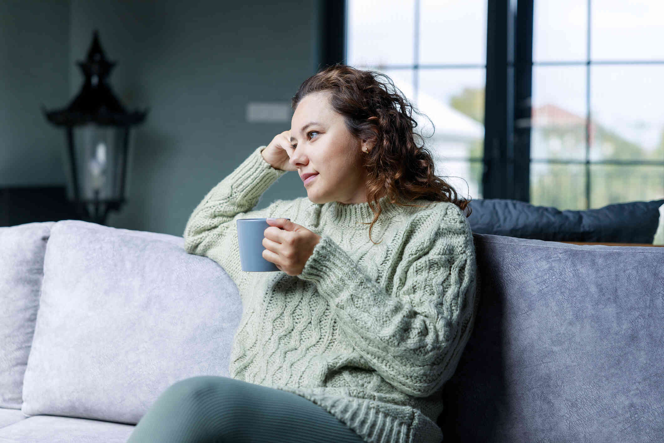 A woman wearing a thick, green sweater sits on a coach while holding a coffee mug and stares our towards the left with a neutral expression.